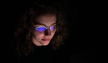 Geceleri bilgisayarıyla çalışan mutlu bir kadın. Oda karanlık ve ekranı gözlüklerine yansıyor..