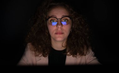 Geceleri bilgisayarla çalışan bir kadın ve gözlükleriyle ekranın yansıması. Kadın yorgun, aşırı çalışma konsepti.