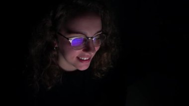 4K, genç bayan geceleri bilgisayarını kullanırken gülümsüyor. Karanlık ve ekran gözlüklerine yansıyor..