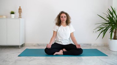 Yatak odasındaki mavi paspasın üzerinde yoga nilüfer pozisyonu alan bir kadın. Kadının sarı kıvırcık saçları var..