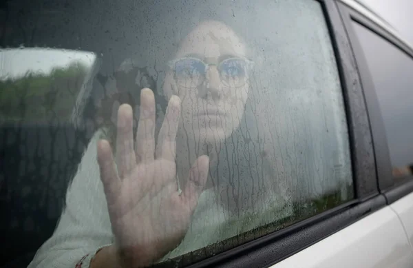 Sad and desolate woman places her hand on the car glass while it is raining. Concept of depression in the car.