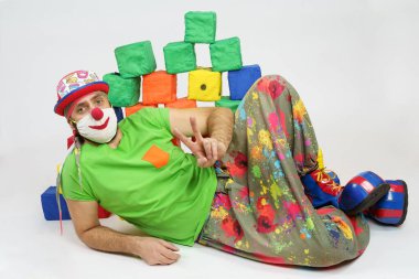 Tatil ve eğlence konsepti. Palyaço yerde yatıyor, arkasında renkli küplerden bir duvar var. Beyazda izole edilmiş