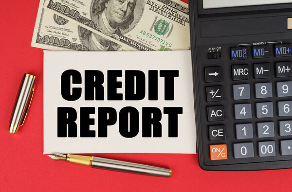 Концепция бизнеса и финансов. На красном фоне, среди денег, калькулятор и ручка лежит знак с текстом - CREDIT REPORT