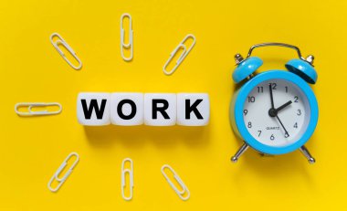 Finance and economics concept. On a yellow background, a blue alarm clock, paper clips and white cubes on which the text is written - WORK