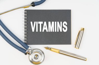 İlaç ve sağlık. Beyaz arka planda bir steteskop, bir kalem ve üzerinde VITAMINS yazan bir defter bulunur.
