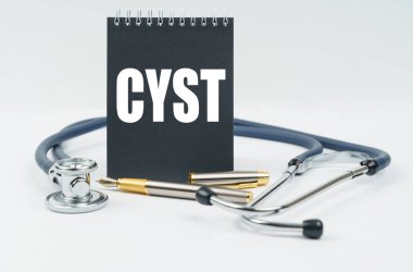 Tıp ve sağlık konsepti. Beyaz arka planda bir stetoskop, bir kalem ve üzerinde CYST yazan siyah bir defter bulunur.