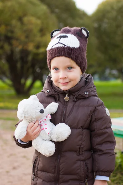 Menino de casaco marrom e chapéu de urso de malha com urso de brinquedo em suas mãos ao ar livre no outono — Fotografia de Stock