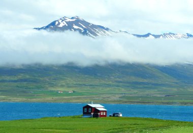 Karlı, sisli dağ ve mavi okyanusa bakan İzlanda 'daki Halka Yolu boyunca uzanan küçük bir evin uzaklık manzarası.