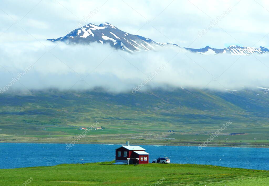The distance view of a small house overlooking the snowy and foggy mountain and blue ocean in the summer along the Ring Road in Iceland