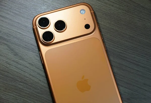 Florida, U.S.A - 2 Ocak 2026 - Ahşap üzerinde üç kameralı turuncu-altın iPhone sergilenen, parlak tasarım ve Apple markalaştırma.