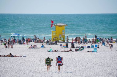 Siesta Key Beach, Florida, ABD - 3 Ocak 2026 - Kırmızı bayraklı parlak sarı cankurtaran kulesi şemsiyeler, güneşlenenler ve okyanus aktiviteleriyle dolu hareketli bir plaj sahnesini denetliyor