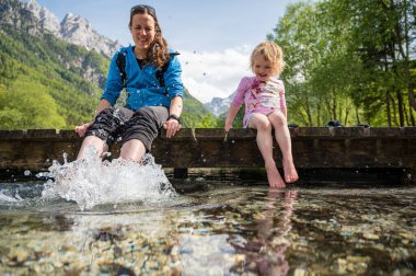 Anne ve kızı ahşap iskelede oturmuş ayaklarıyla su sıçratarak eğleniyorlar..