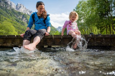 Anne ve kızı ahşap iskelede oturmuş ayaklarıyla su sıçratarak eğleniyorlar..
