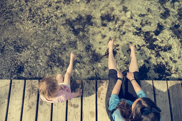 Vue plongeante de la mère et de la fille assises sur un quai en bois avec les pieds dans l'eau. — Photo