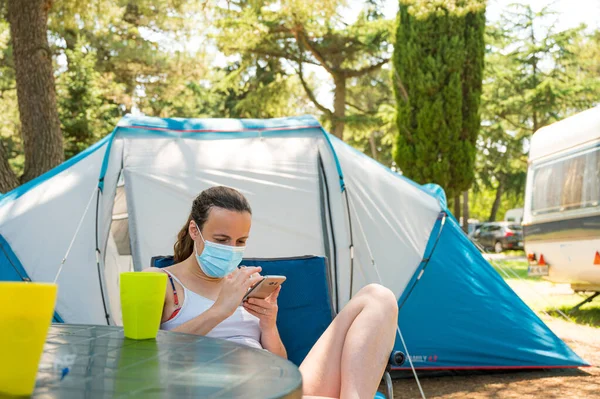 Kamp çadırının önünde maske takan bir kadın akıllı telefonunu kontrol ediyor.. Stok Resim