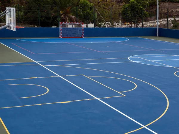 A blue and white large playing field as a sports field outdoors in nature for all ball games such as basketball, volleyball, dodgeball, four corners football, a net goal can be seen from the front, the floor is painted blue with various white play li