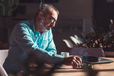 Orta yaşlı sakallı bir adam sabah kahvesini içip bir kafede sigara içiyor. İş adamları
