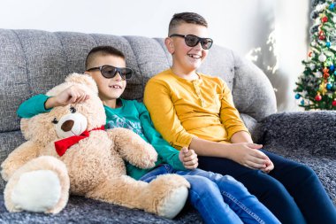 Sevimli çocuklar, 3 boyutlu gözlüklü film izlerler oturma odasındaki kanepede oyuncak ayıyla otururlar.