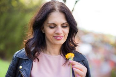 Siyah deri ceketli güzel, orta yaşlı bir kadın gülümsüyor, hayattan zevk alıyor, sarı çiçek kokuyor. Açık havada.