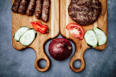 Dilimlenmiş salatalıklı, domatesli, soğanlı, kırmızı biberli, ızgara etli, Türk hamburgerli ahşap mutfak tahtası.