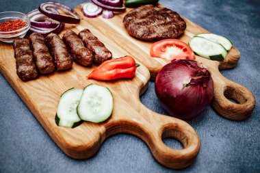 Ahşap mutfak tahtasıyla ızgara et, Türk hamburgeri ve dilimlenmiş salatalık, domates, soğan ve kırmızı biber.