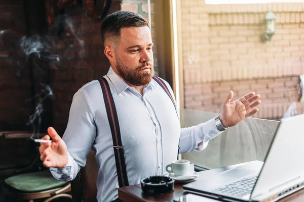 Genç hippi işadamı bir kafede, sigara içerken ve kahve içerken bir video görüşmesi yapıyor.