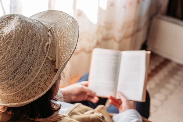 Elderly woman with a hat on, reading a book by the window. Afternoon light