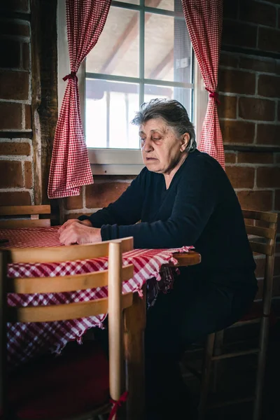 おばあちゃんは家の窓のそばに一人で座って悲しく心配していた — ストック写真