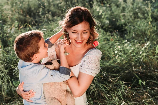 美しい若いお母さんは彼女の腕の中で彼女のブロンドの息子を保持する 彼は彼女の髪に花を入れている 屋外の写真 母の日 — ストック写真