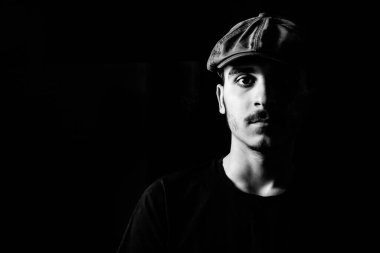 Siyah beyaz bıyıklı, siyah arka planda şapkası olan genç bir adamın portresi..