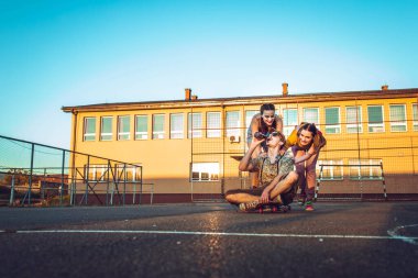 Bir grup arkadaş dışarıda takılıyor. İki kız, erkek arkadaşını kaykay tahtasına itiyor.