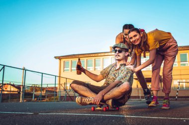 Bir grup arkadaş dışarıda takılıyor. İki kız, erkek arkadaşını kaykay tahtasına itiyor.