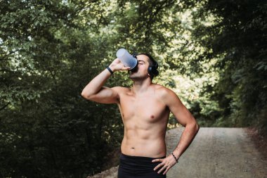 Genç çıplak göğüs adam açık havada su içiyor. Koşu, spor aktivitesi