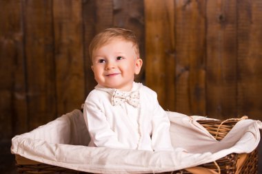 baby blond boy in a white suit white socks sitting in basket on a wooden background. next to a small duckling