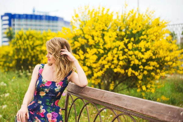 Una hermosa joven rubia sentada en un banco del parque en un vestido colorido en colores brillantes — Foto de Stock