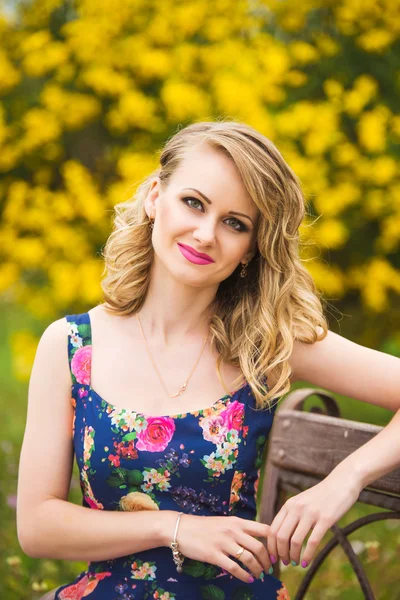 Uma bela jovem loira sentada em um banco de parque em um vestido colorido em cores brilhantes — Fotografia de Stock