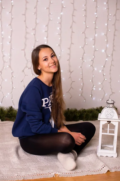 Beautiful young girl in a sweatshirt and tights near the Christmas tree, Christmas lights in the background, she smiles, happy, looking directly — Stock Fotó