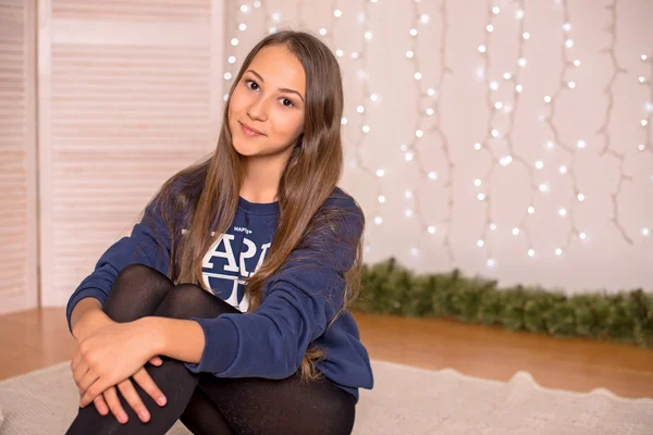Beautiful young girl in a sweatshirt and tights near the Christmas tree, Christmas lights in the background, she smiles, happy, looking directly — Stok fotoğraf