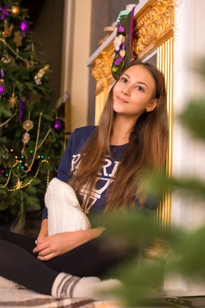 Beautiful young girl in a sweatshirt and tights near the Christmas tree, Christmas lights in the background, she smiles, happy, looking directly — Stockfoto