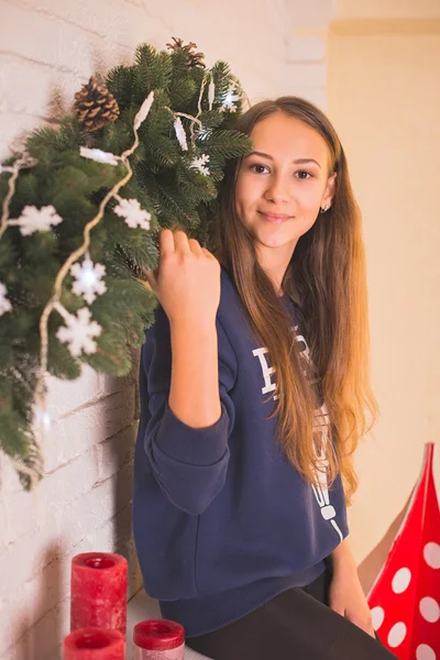 Schönes junges Mädchen in Sweatshirt und Strumpfhose in der Nähe des Weihnachtsbaums, Weihnachtsbeleuchtung im Hintergrund, sie lächelt, glücklich, schaut direkt — Stockfoto