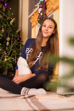 beautiful young girl in a sweatshirt and tights near the Christmas tree, Christmas lights in the background, she smiles, happy, looking directly,white socks, warm socks, a white teddy bear, hugging a pillow,