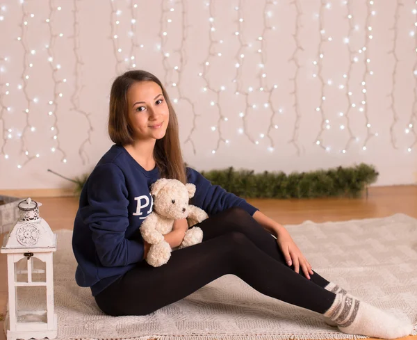 Menina bonita em uma camisola e meia perto da árvore de Natal, luzes de Natal no fundo, ela sorri, feliz, olhando diretamente, meias brancas, meias quentes, um urso de pelúcia branco, abraçando um travesseiro , — Fotografia de Stock