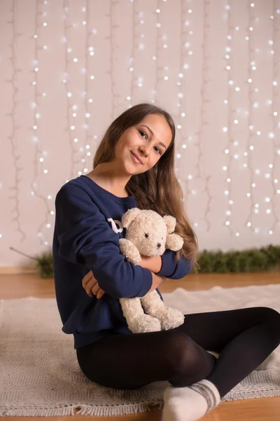 Beautiful young girl in a sweatshirt and tights near the Christmas tree, Christmas lights in the background, she smiles, happy, looking directly,white socks, warm socks, a white teddy bear, hugging a pillow, — Stock fotografie