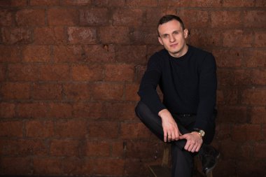 wealthy man, dark background, short-haired man, businessma, a man in a dark  sweater, blazer, looking directly into the camera, stern look, business, brick wall, dark background, red brick, bright smile,