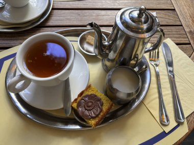 Bir fincan çay, metal çaydanlık, süt sürahisi ve bir kafede tepside sunulan küçük bir pasta ile sıcak ve rahatlatıcı bir sabah atmosferini çağrıştırıyor.