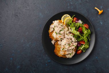 Kızarmış morina, patatesli krep, mantar sosu ve salata ile servis edilir. Siyah bir tabakta porsiyon. Koyu arkaplan.