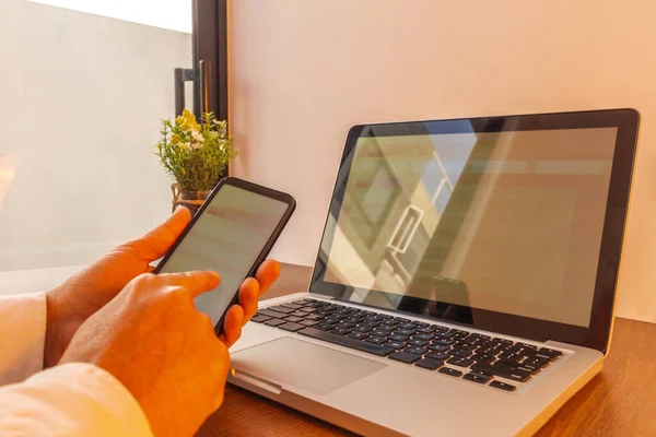 hands of a man navigating through his cell phone with a laptop computer coordinated with the same website. Image in warm tones on a wooden desk in an office