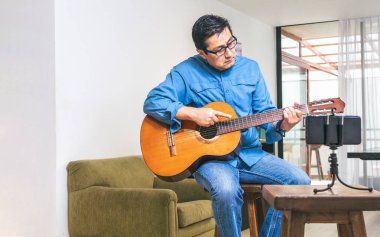 Gitar öğretmeni online bir derste gitar akorunu gösteriyor. Cep telefonu oturma odasındaki bankta oturuyor.