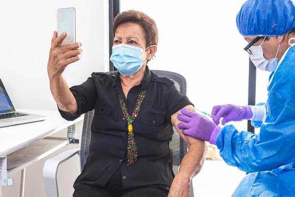 Yaşlı yetişkin bir kadın, salgın sırasında bir doktordan koronavirüs aşısı alırken cep telefonuyla selfie çekiyor. Teknoloji ve aşı kampanyası konsepti