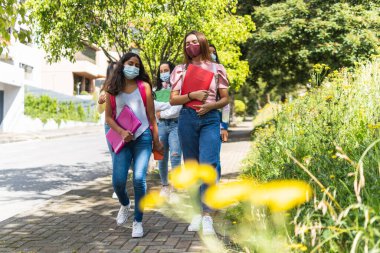 Liseye maskeli ve diğer sınıf arkadaşlarıyla birlikte sırt çantaları ve not defterleriyle odak dışı giden iki arkadaşın seçici fotoğrafıyla yakın çekim.